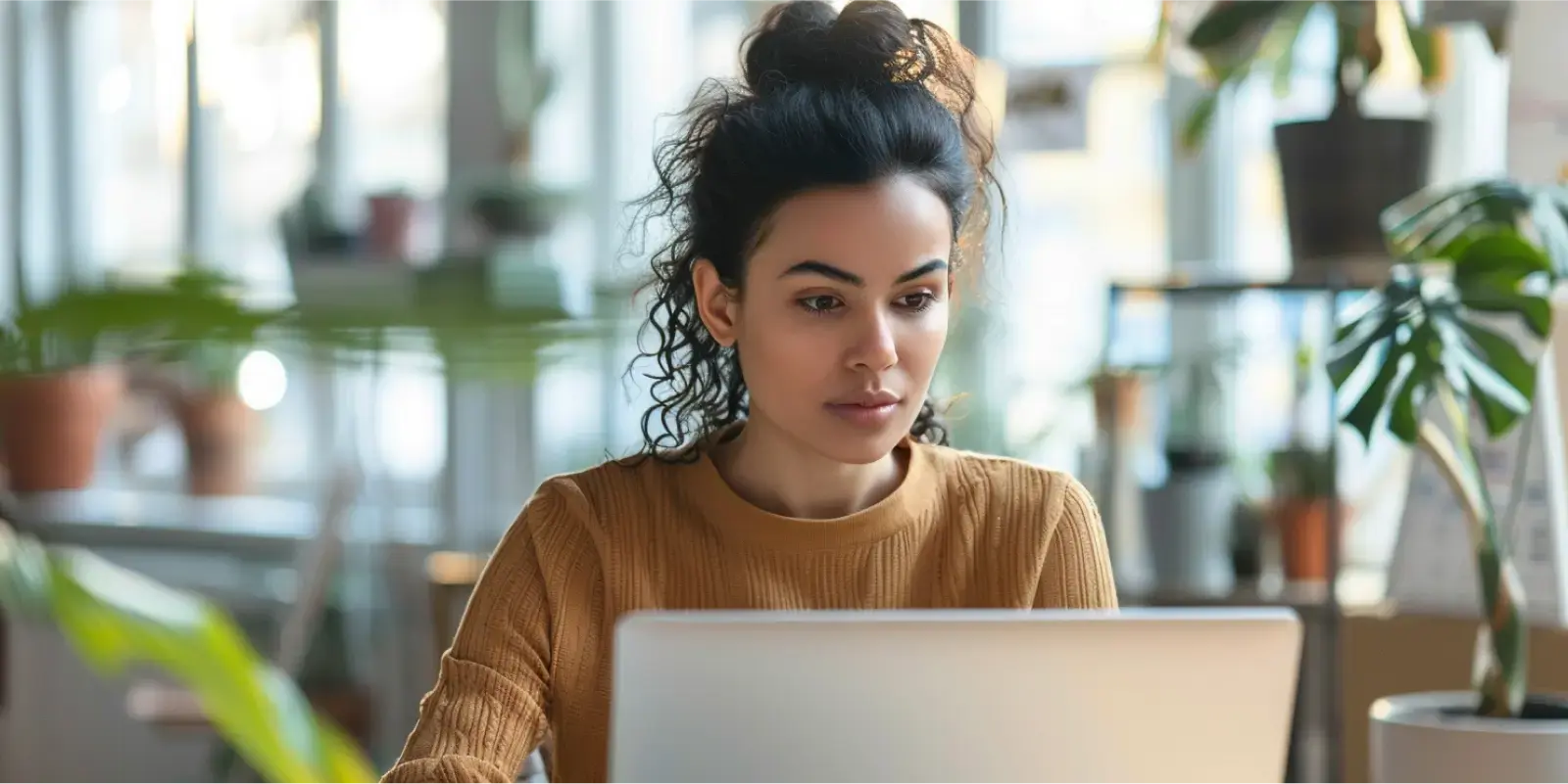 Mulher concentrada trabalhando em um laptop lendo sobre follow up em um ambiente de escritório iluminado e com plantas ao fundo