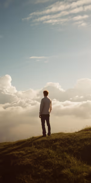 Person standing on a grassy hilltop overlooking a sea of clouds under a blue sky.