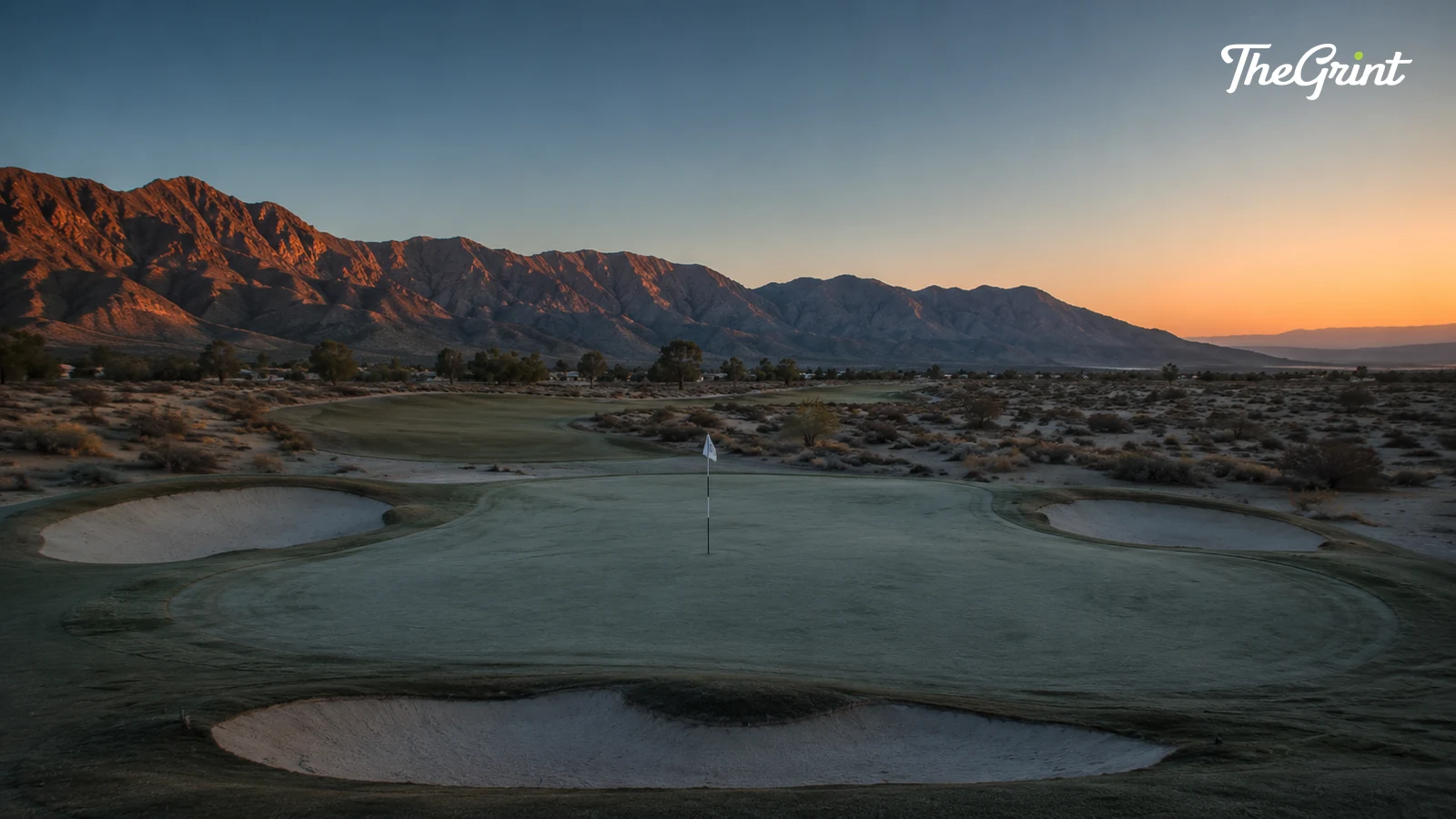 Desert golf green with bunkers and a flagstick below mountains at sunset