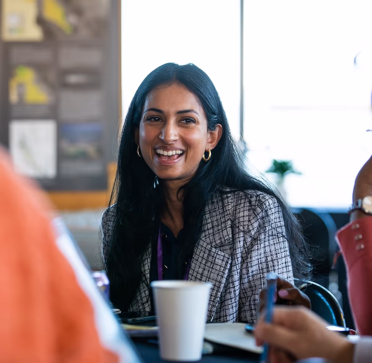 A woman in a checkered blazer smiles brightly while seated at a table during a group discussion. Coffee cups, notebooks, and blurred participants are visible in the foreground.