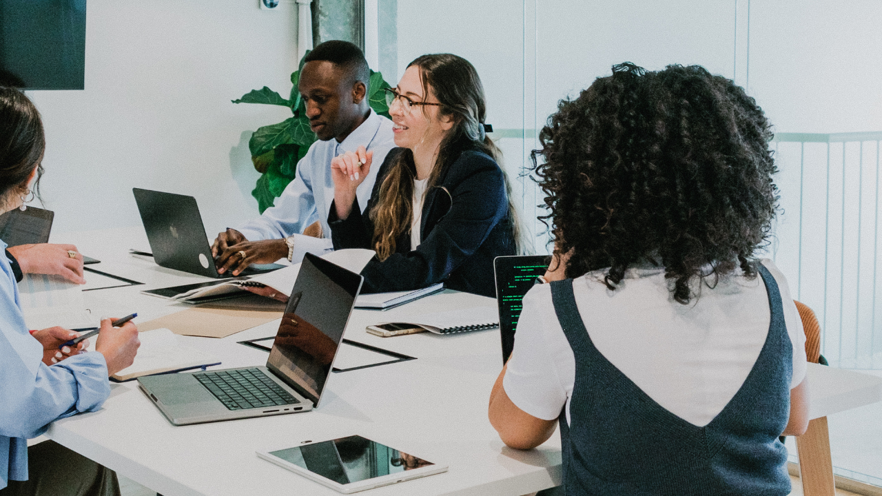 A group of professionals in discussion around a table with laptops and notebooks, collaborating in a bright, modern office space.