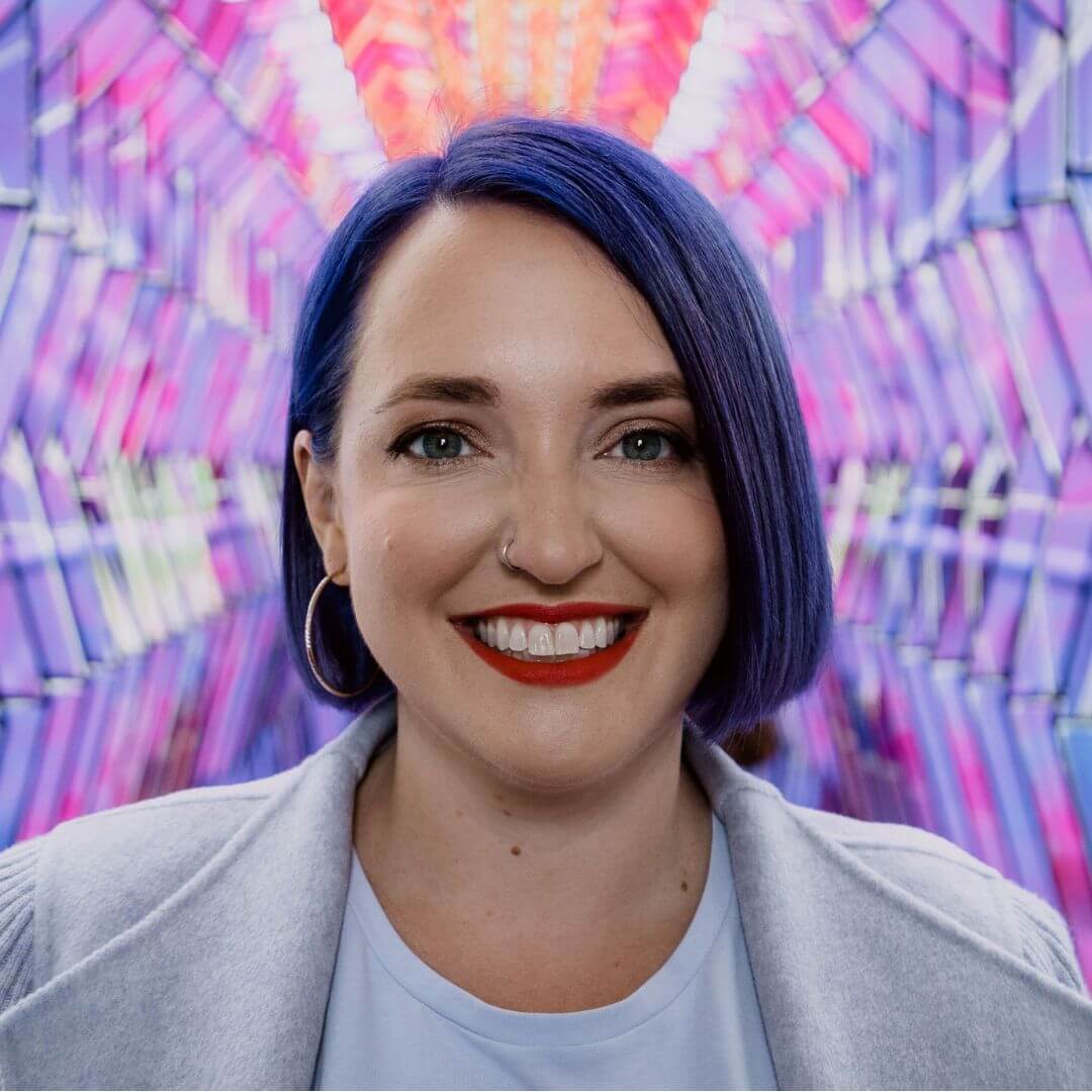 Headshot of Aubrey Blanche smiling with dark hair and red lipstick, wearing a light blue top and jacket, standing in front of a colourful geometric background.