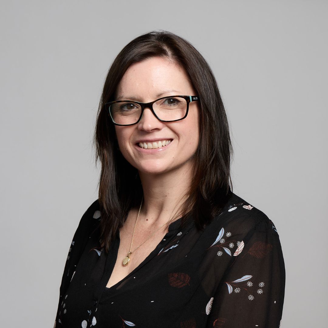 Headshot of Amy Whalley smiling with straight dark hair and glasses, wearing a black patterned top against a light grey background.