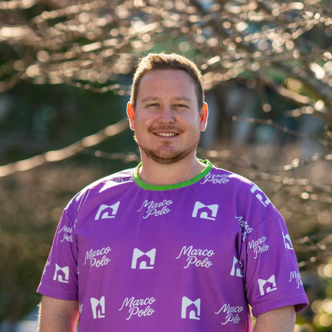 Headshot of Aaron Cotton smiling with short light brown hair, wearing a purple Marco Polo shirt with white logos, standing outdoors in natural light.