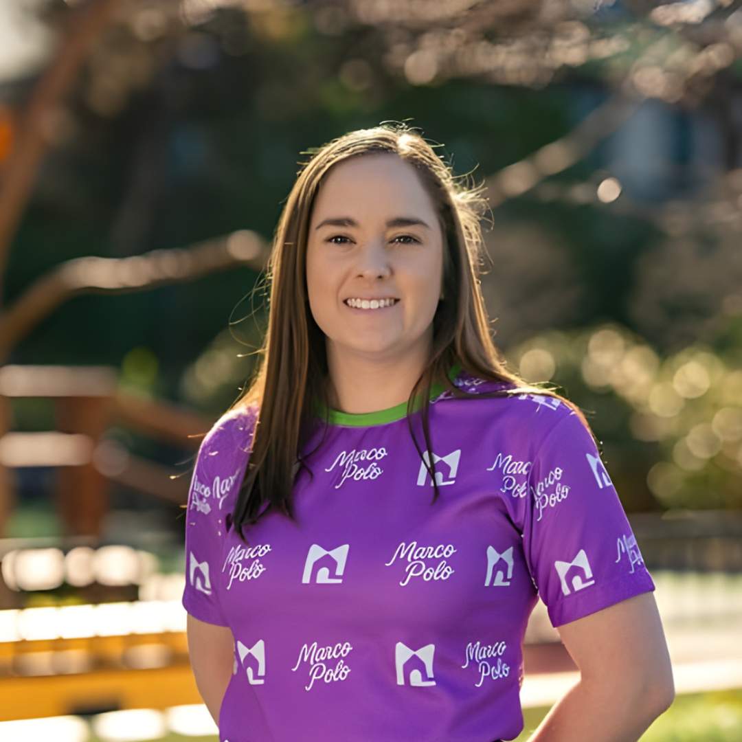 Headshot of Amy Connelly smiling with straight brown hair, wearing a purple Marco Polo shirt with white logos, standing outdoors in natural light.