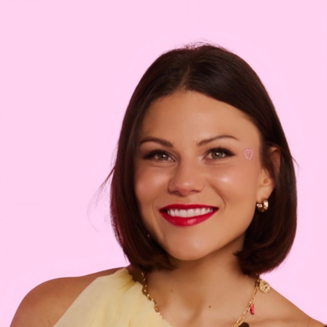 Headshot of Storm Menzies smiling with short dark hair and red lipstick, wearing gold jewellery and a pale yellow top against a pink background.