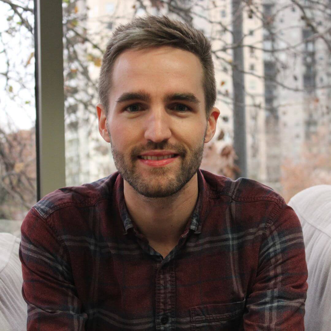 Headshot of Adam Jahnke smiling while seated indoors, wearing a dark red plaid shirt. A window behind Adam shows trees and blurred buildings in the background.