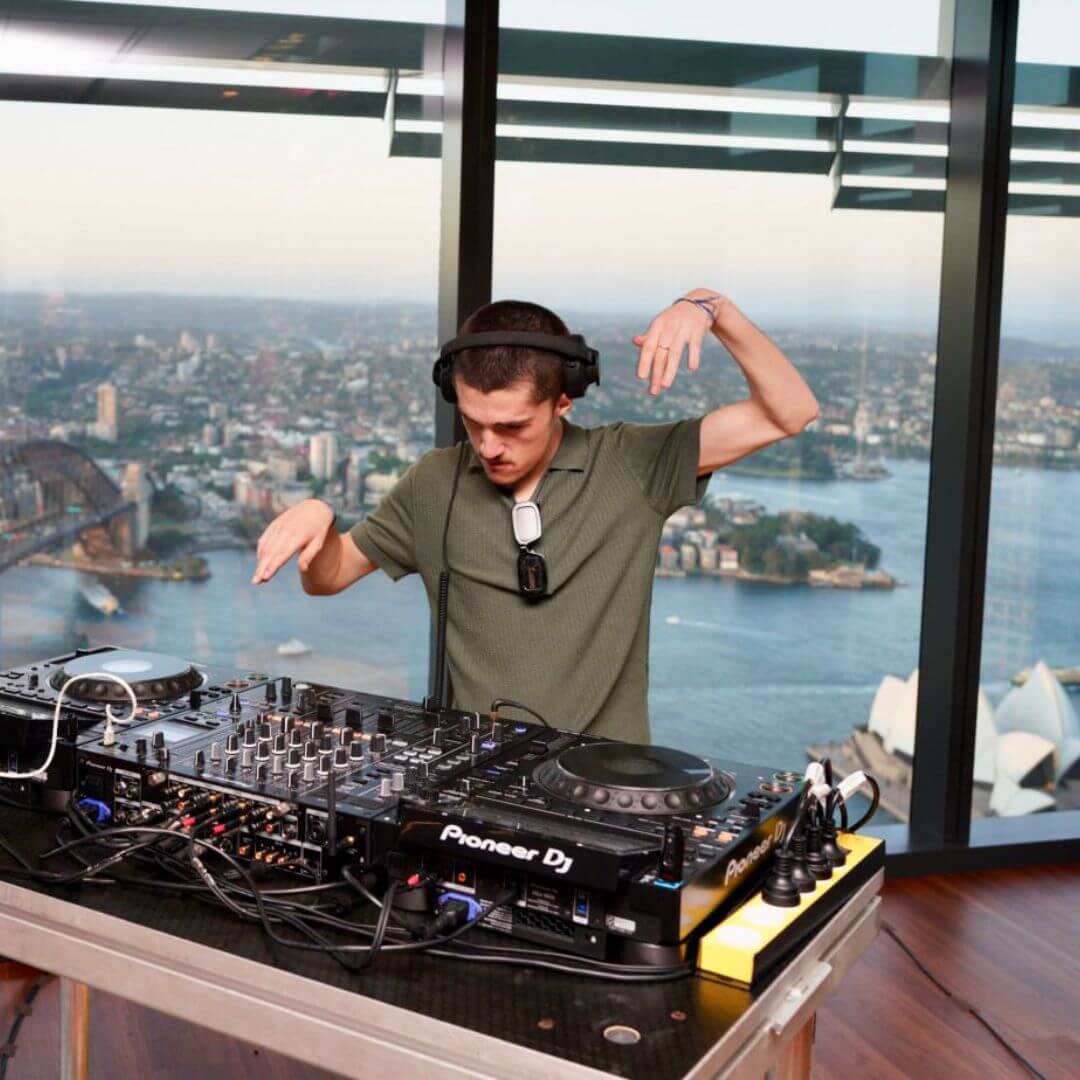Person wearing headphones and a green shirt performing as a DJ with Pioneer DJ equipment, with a city and harbour view visible through large windows behind them.