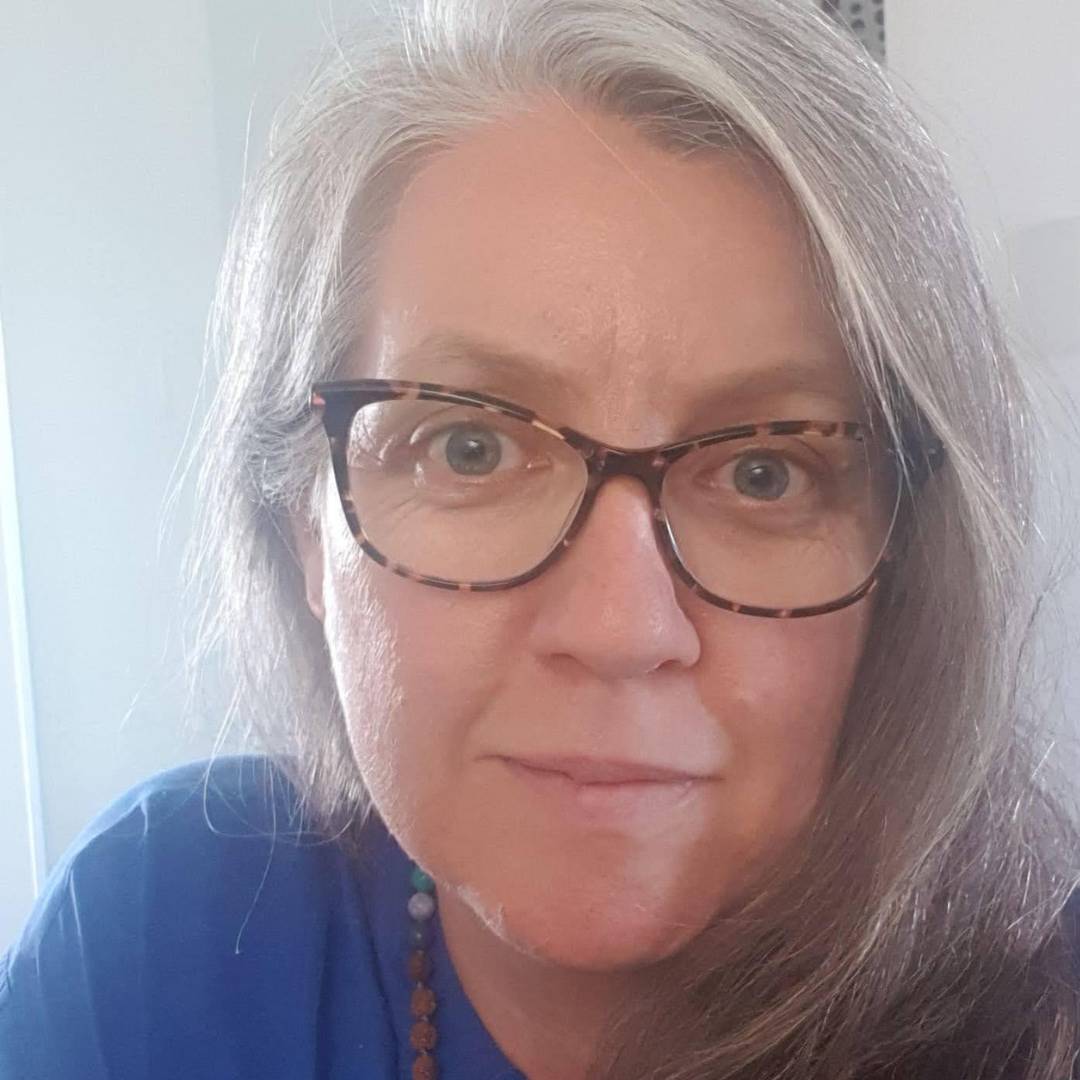 Headshot of Jade Grimwood smiling softly, wearing glasses, a blue top, and a beaded necklace, with long grey hair over one shoulder against a softly lit indoor background.