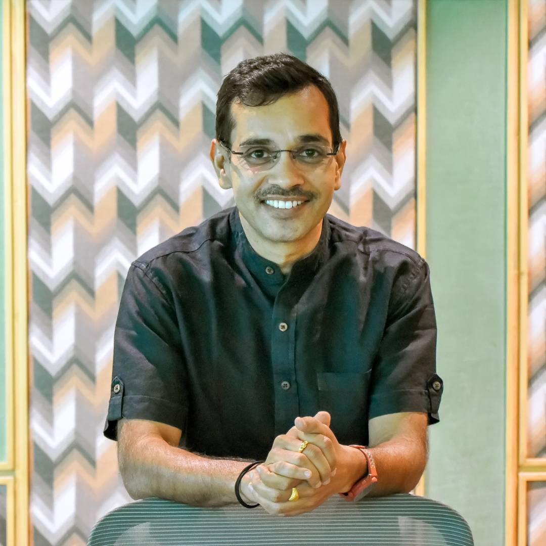 Hadshot of Prateek Madhav smiling, wearing glasses and a dark short-sleeved shirt, leaning forward with hands clasped in front of a patterned background with chevron designs in green, white, and orange tones.