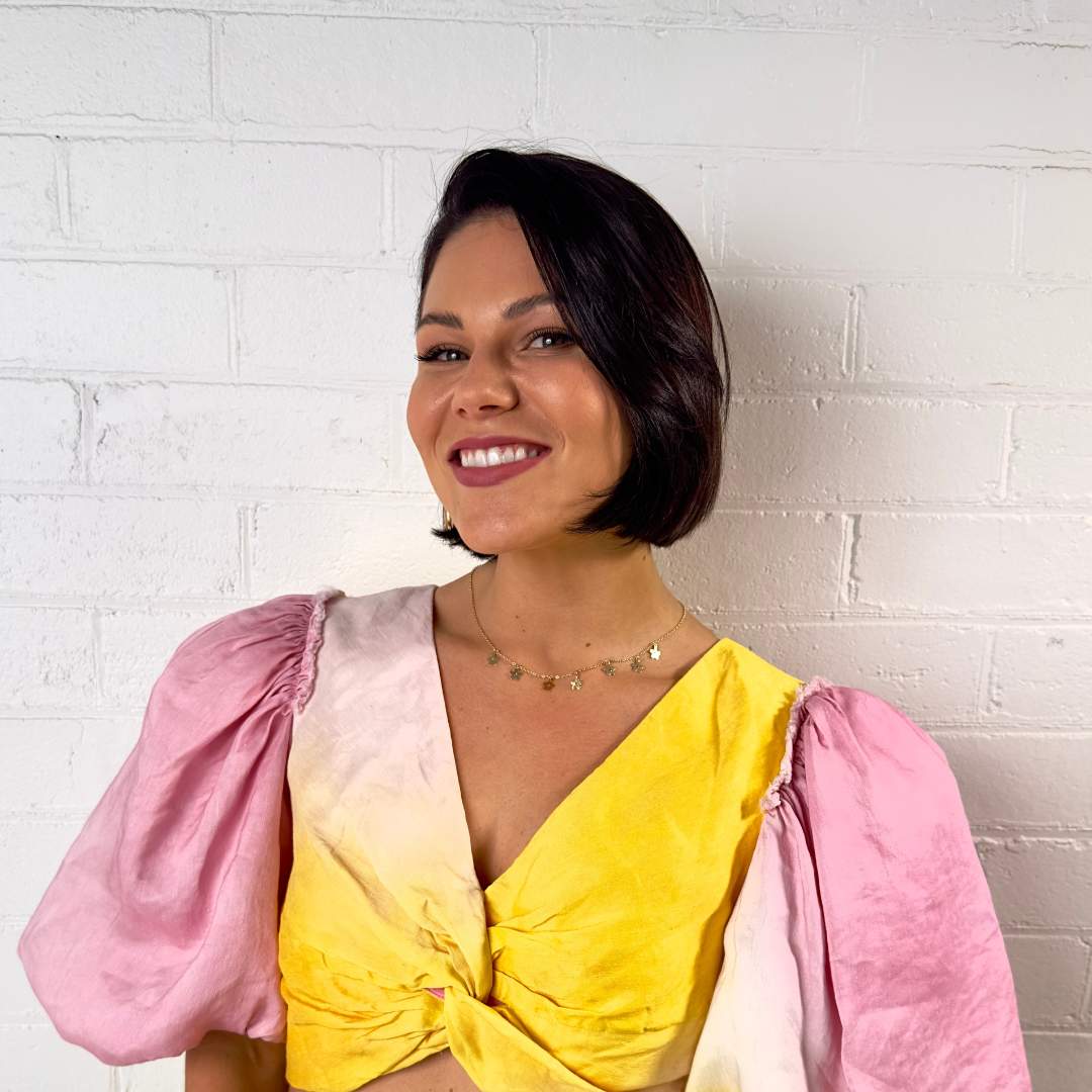 Headshot of Storm Menzies smiling, wearing a yellow and pink puff-sleeved top and a delicate gold necklace, standing against a white brick wall background.