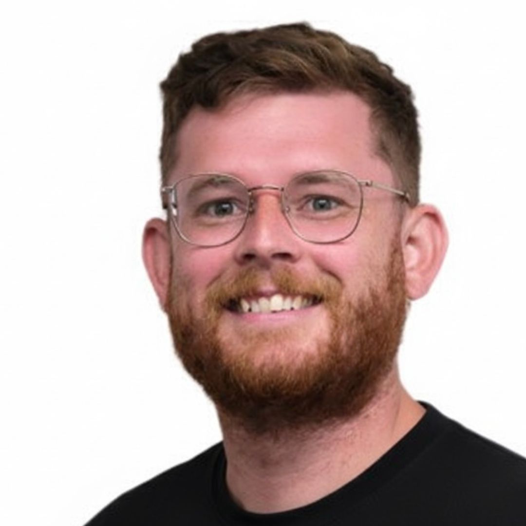 Alex Jones smiling with short brown hair, beard, and glasses, wearing a black shirt against a plain white background.