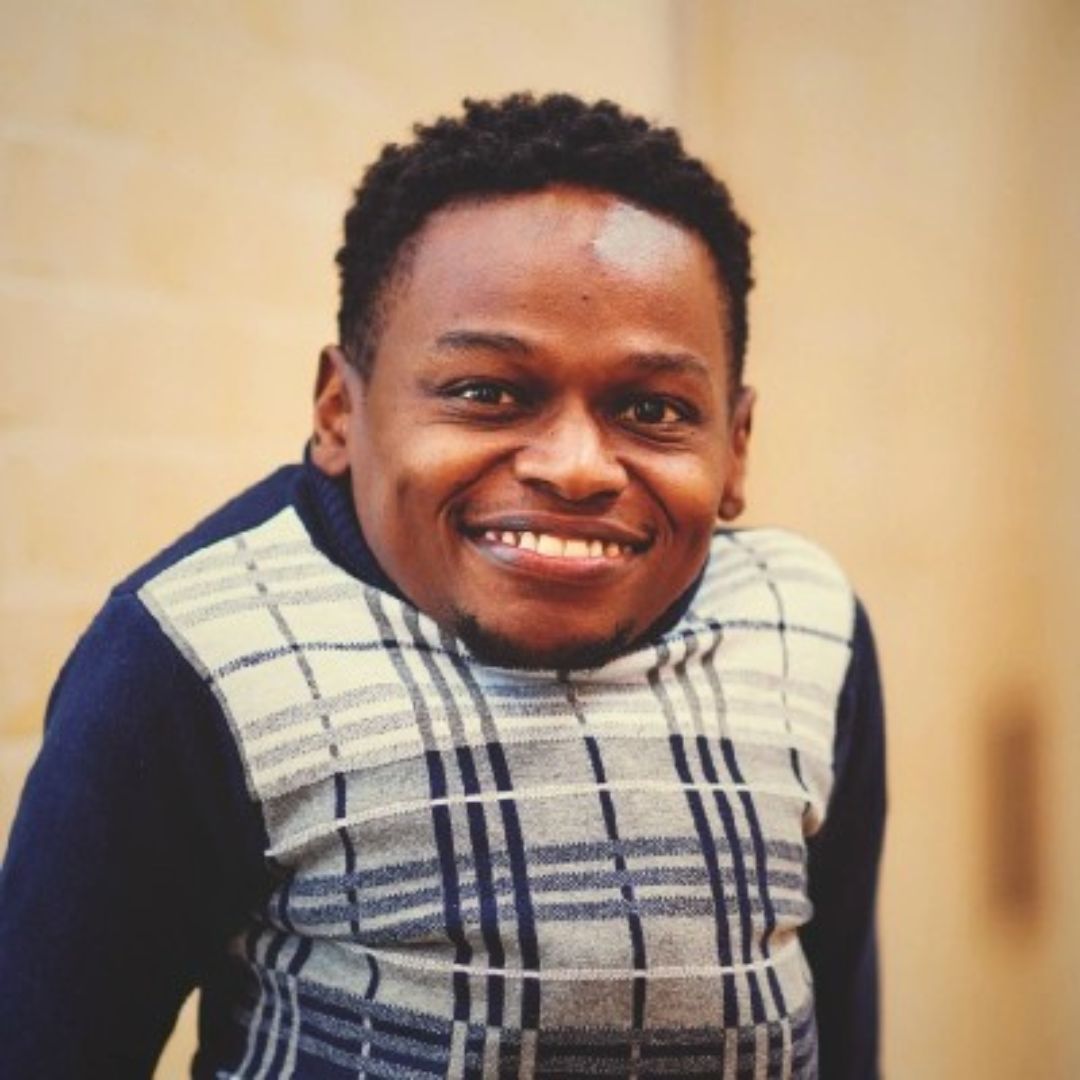 Bernard Chiira wearing a navy and cream checkered sweater, standing against a light tan background.