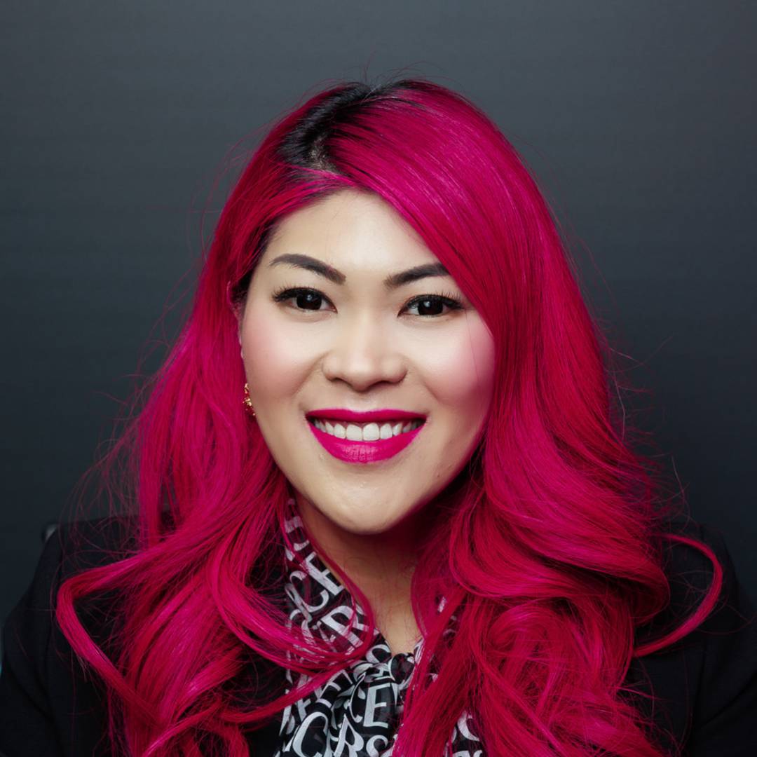 Daisy Wong smiling with bright pink hair, wearing a patterned blouse and dark blazer against a dark grey background.