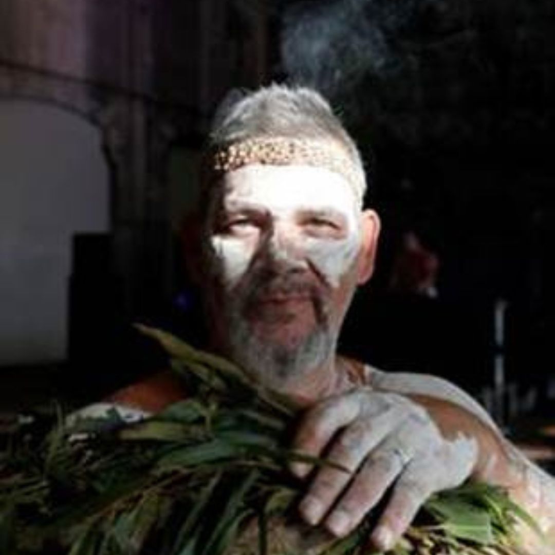 Matthew Doyle with traditional white body paint and a woven headband, holding green eucalyptus leaves during a cultural ceremony.