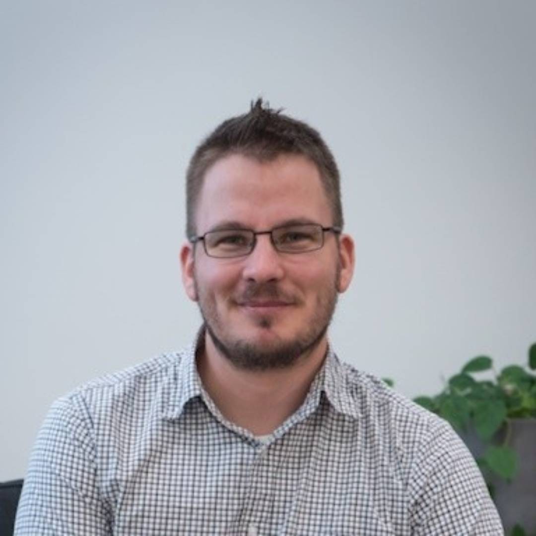 Steve King smiling with short hair and glasses, wearing a checkered shirt, seated indoors with a plant in the background.