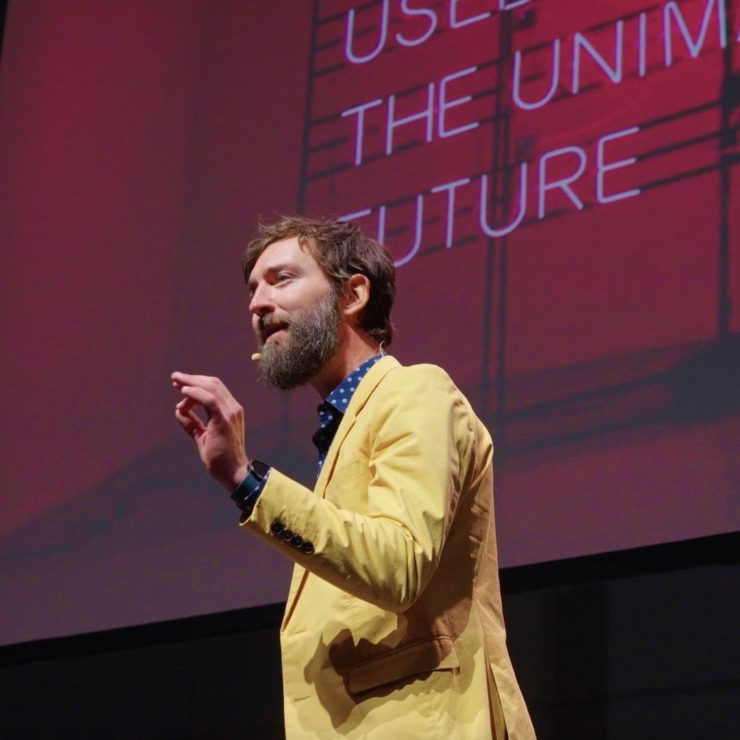 Tāne Hunter with a beard speaking on stage, wearing a yellow jacket and headset microphone, with a red presentation screen behind him displaying the words “Future Crunch.”