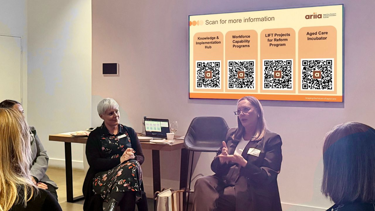 A small group sits in discussion at an ARIIA event. Two presenters are seated at the front, one gesturing while speaking, with a presentation screen behind them displaying QR codes for ARIIA programs.