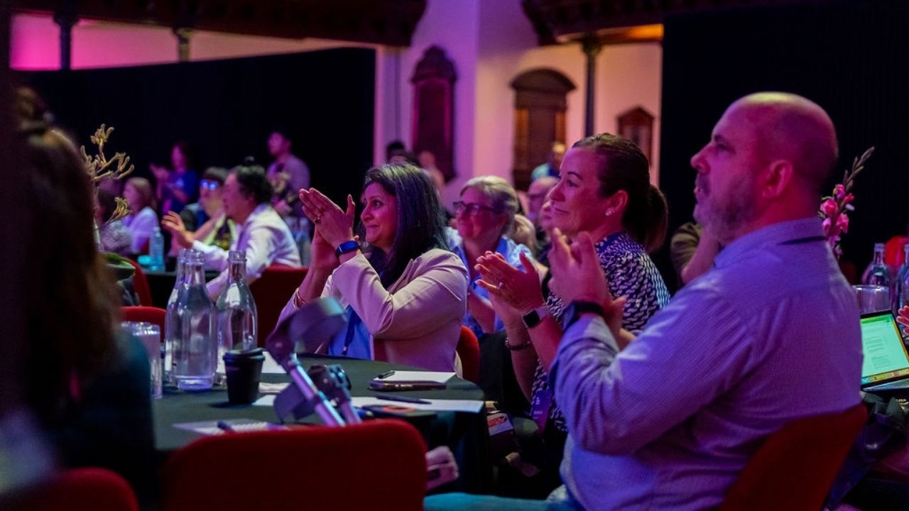 Audience members seated at round tables applaud during a Disability Tech Summit 2025 session, illuminated by purple and blue stage lighting. The room is full, with people smiling and engaged as they watch the presentation.