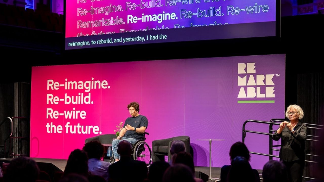 A keynote presenter using a wheelchair presents on stage at the Disability Tech Summit 2025, seated in front of a large pink and purple backdrop that reads “Re-imagine. Re-build. Re-wire the future.” A sign language interpreter stands to the right, and live captions are displayed on a screen above the stage.