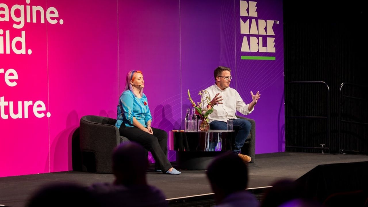 Two presenters sit on stage in conversation at the Disability Tech Summit 2025, framed by a large pink and purple backdrop reading “Re-imagine. Re-build. Re-wire the future.” The woman on the left, wearing a bright blue top, listens attentively, while the man on the right gestures as he speaks. A small table with water bottles and flowers sits between them, and audience members are visible in the foreground.