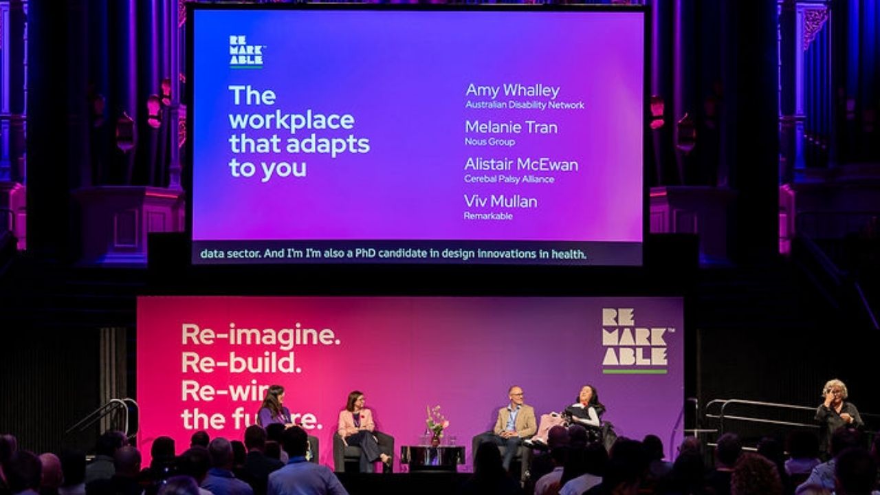 A panel discussion takes place on stage at the Disability Tech Summit 2025 beneath a large screen titled “The workplace that adapts to you,” listing speakers Amy Whalley, Melanie Tran, Alistair McEwan, and Viv Mullan. Four panellists sit in armchairs on a pink and purple backdrop reading “Re-imagine. Re-build. Re-wire the future.” A sign language interpreter stands to the right, and audience members are visible in the foreground.