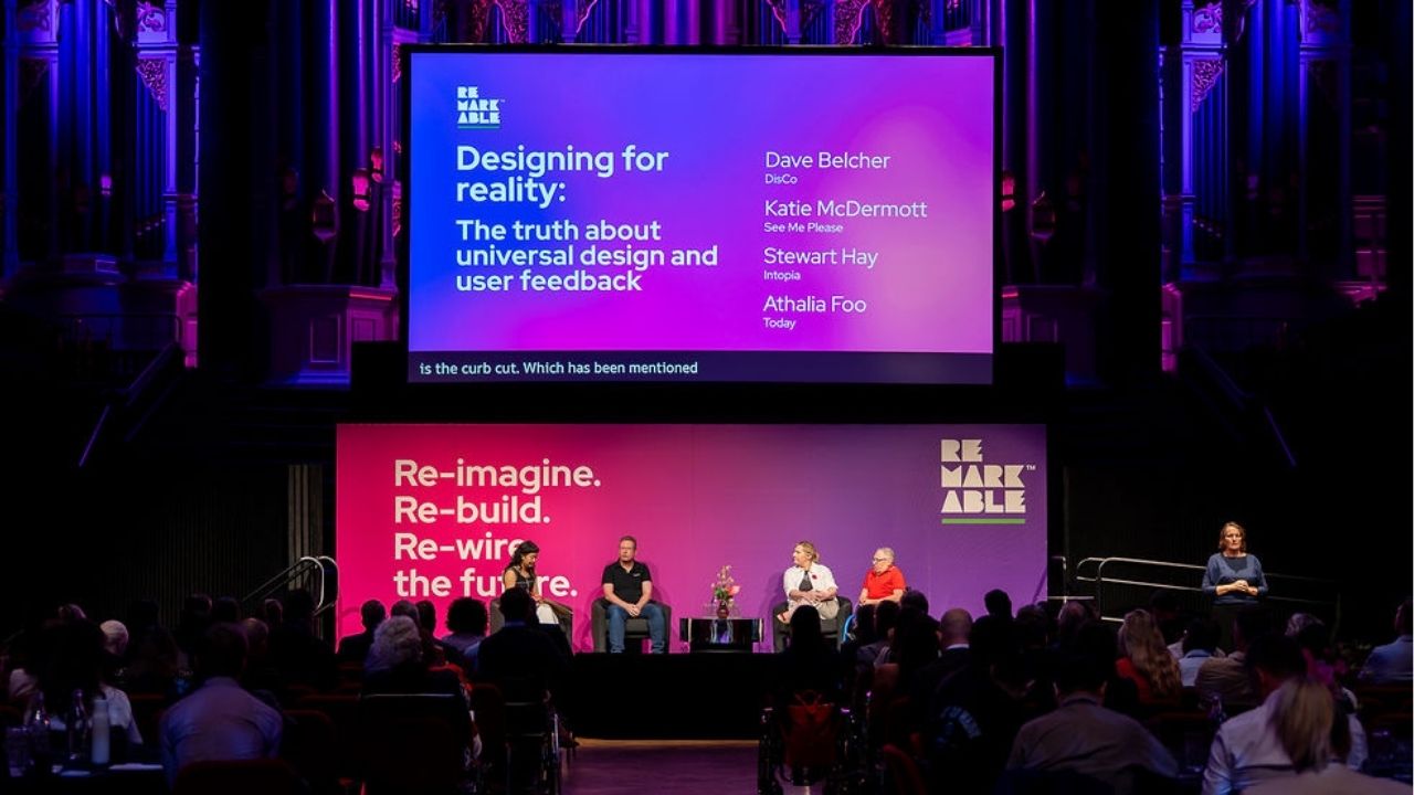 A panel discussion takes place on stage at the Disability Tech Summit 2025 beneath a large screen titled “Designing for reality: The truth about universal design and user feedback,” listing speakers Dave Belcher, Katie McDermott, Stewart Hay, and Athalia Foo. Four panellists sit on stage against a pink and purple backdrop reading “Re-imagine. Re-build. Re-wire the future.” A sign language interpreter stands to the right, and the audience is visible in the foreground.