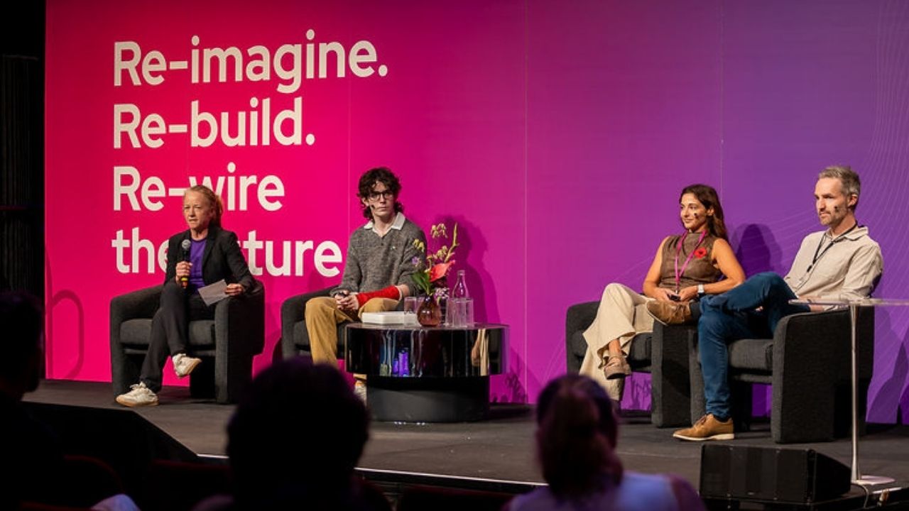 Four presenters sit on stage for a panel discussion at the Disability Tech Summit 2025, framed by a large pink and purple backdrop reading “Re-imagine. Re-build. Re-wire the future.” The moderator on the left holds a microphone, while the three panellists sit beside a small table with flowers and water bottles. Audience members are visible in the foreground.