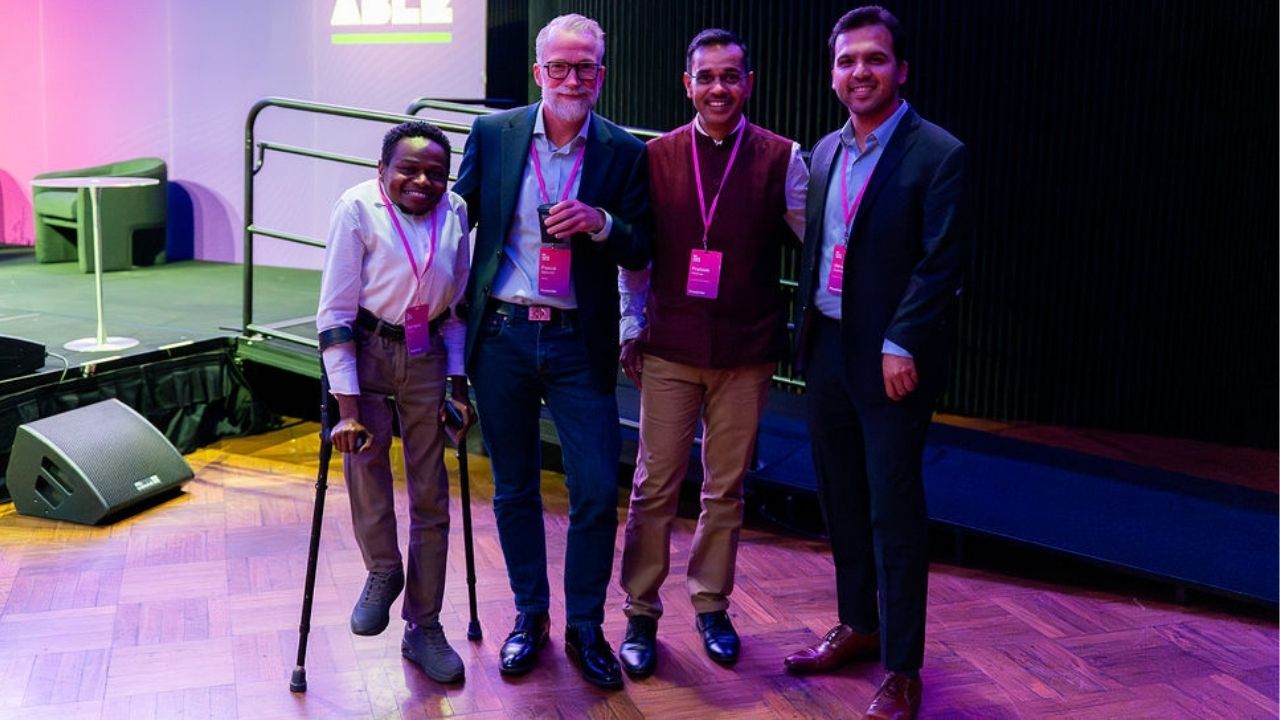 Four presenters stand together smiling for a photo at the Disability Tech Summit 2025. One person uses forearm crutches, and all wear Summit lanyards as they pose in front of the stage area under purple lighting