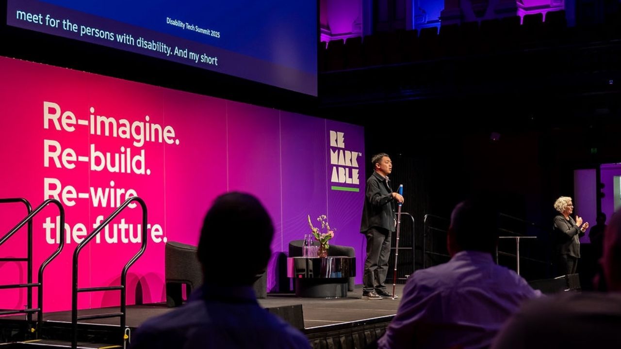 A presenter stands on stage at the Disability Tech Summit 2025 holding a white cane, delivering a presentation in front of a large pink and purple backdrop reading “Re-imagine. Re-build. Re-wire the future.” A sign language interpreter stands to the right, and audience members watch from the foreground.