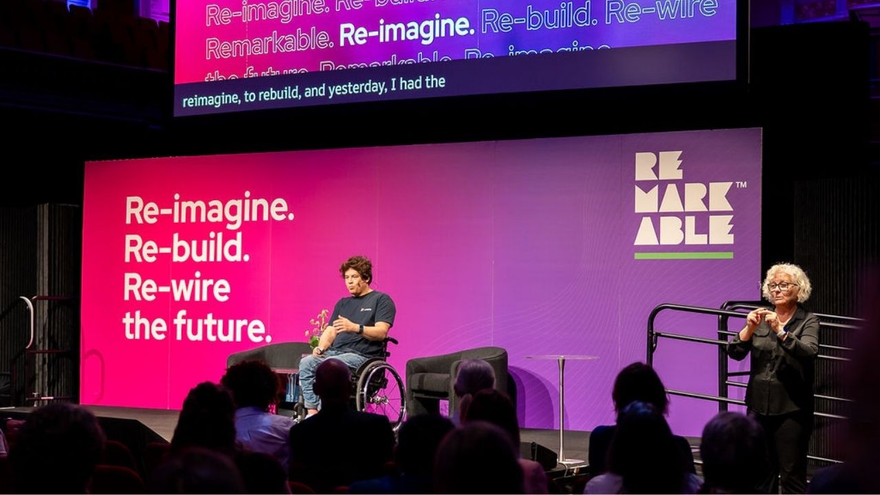 A keynote presenter using a wheelchair presents onstage at the Disability Tech Summit 2025, seated in front of a bright pink and purple backdrop that reads “Re-imagine. Re-build. Re-wire the future.” A sign language interpreter stands to the side, with an audience watching in the foreground.