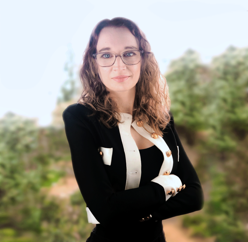 Flic is a caucasian woman with strawberry blonde wavy hair and glasses, she has blue eyes. She is wearing a navy blue jacket with gold buttons and white details, she stand with arms crossed facing the camera smiling and there are trees behind her.