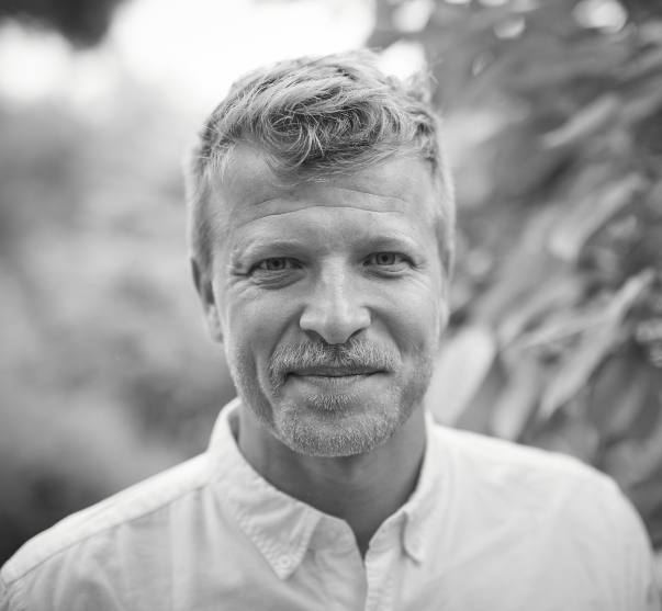 A smiling man wearing a white button-down shirt, standing in front of some trees. 