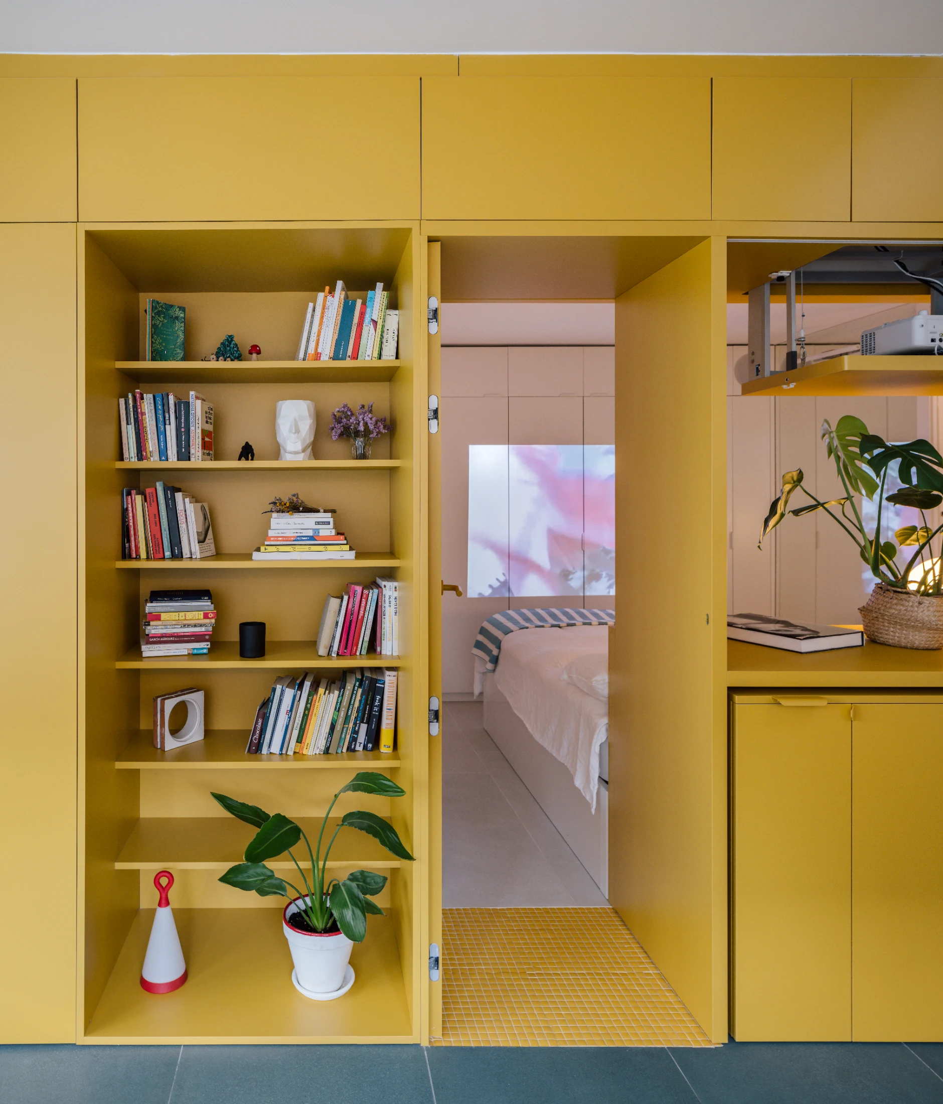 A wall of yellow cabinetry and shelving separates the bedroom from the multifunctional kitchen meals and living space in Casa Gialla (2022). Taking cues from medieval dwellings where