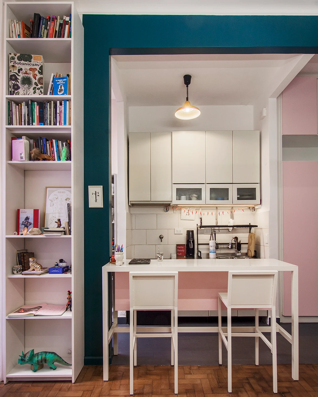The apartment's original compact single-wall kitchen in Billinghurst has been transformed into a practical and social space with the addition of a narrow white and pink breakfast bar. The MoobArq team also built a clever and generous storage space for spic