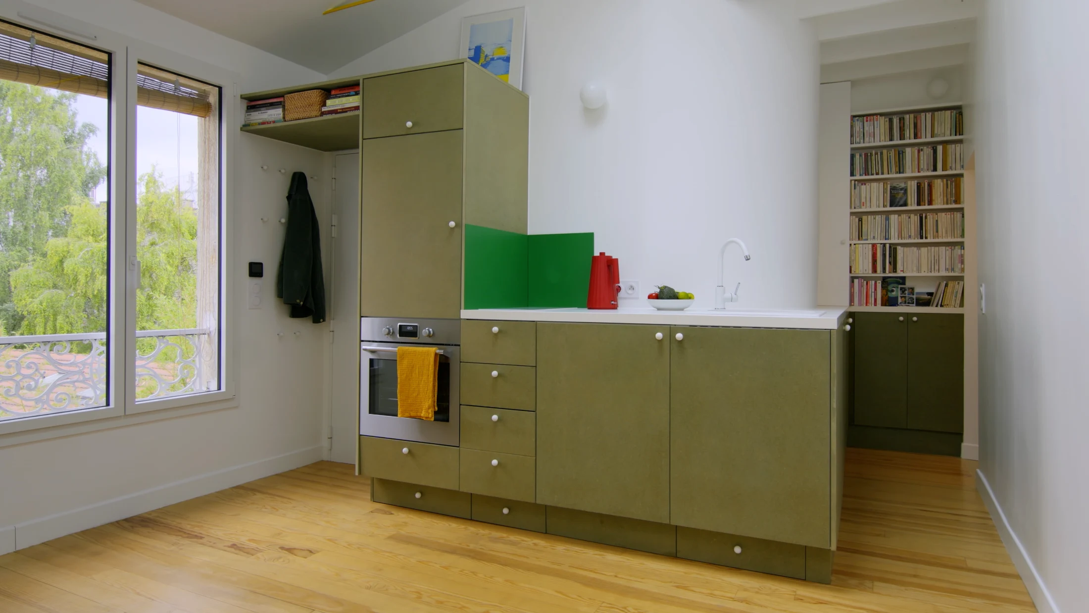 In the corridor of Rue de Bagnolet designed by QSA, the green MDF kitchen cabinets and white laminate worktop continue outside the expected footprint of the kitchen and along the curve of its internal wall, providing additional storage for dinnerware, cook