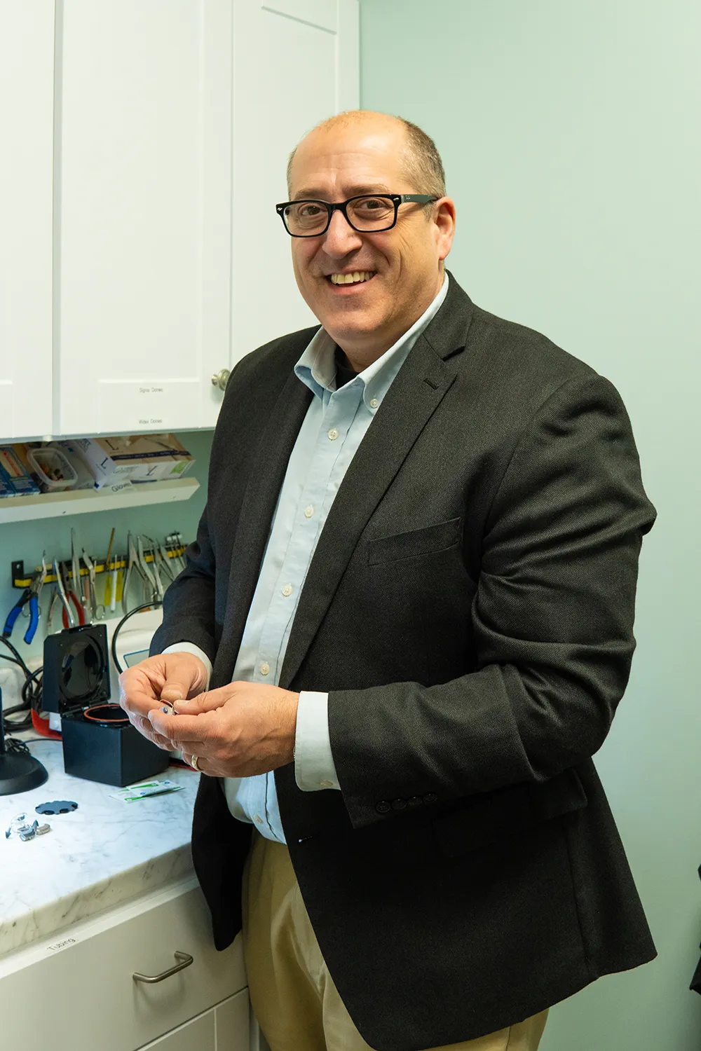 Smiling female audiologist holding glasses stands and talks to an elderly woman seated in an office with plants and paperwork in the background.