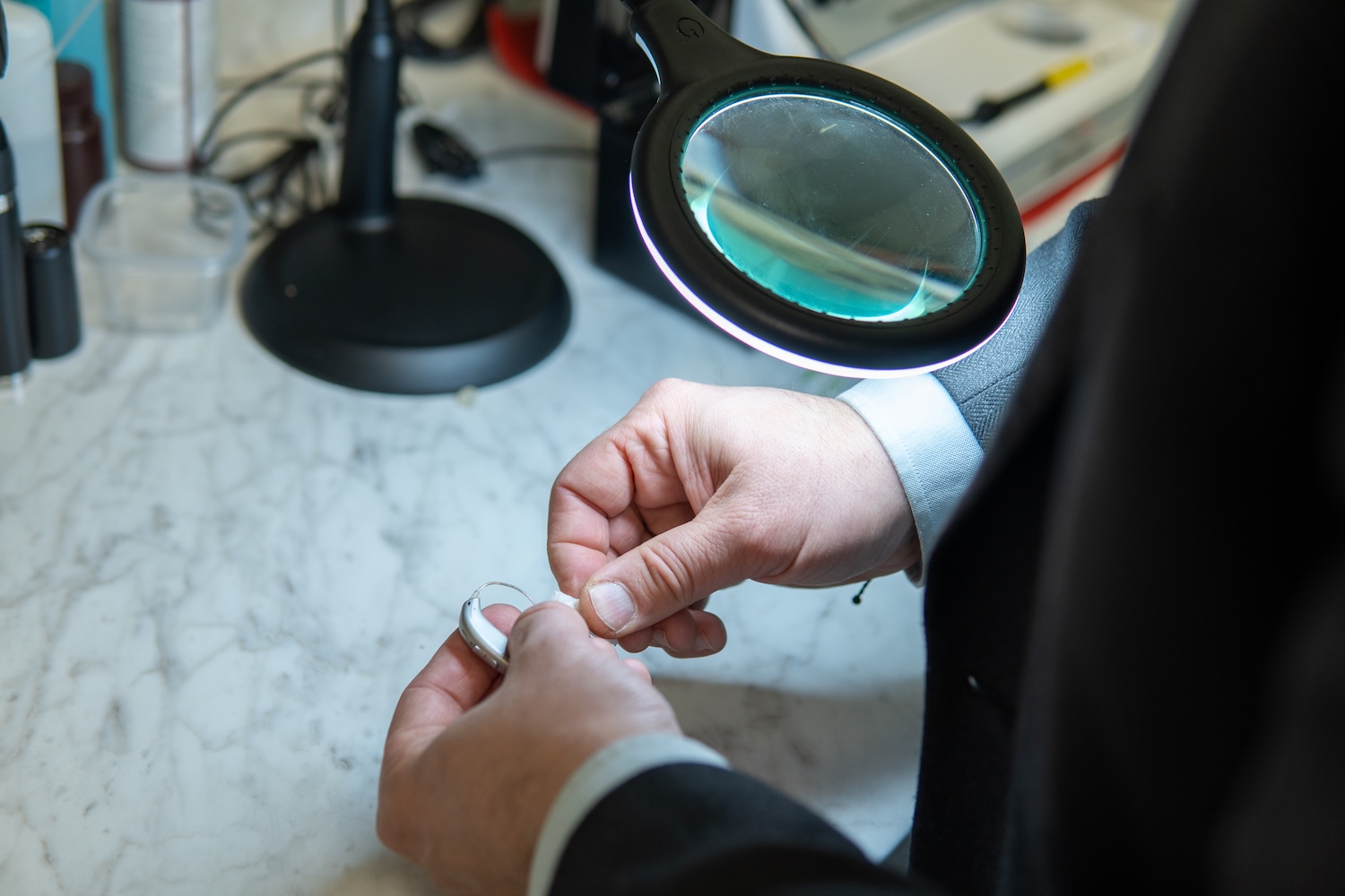 Person holding and inspecting a hearing aid under a magnifying lamp on a marble surface.
