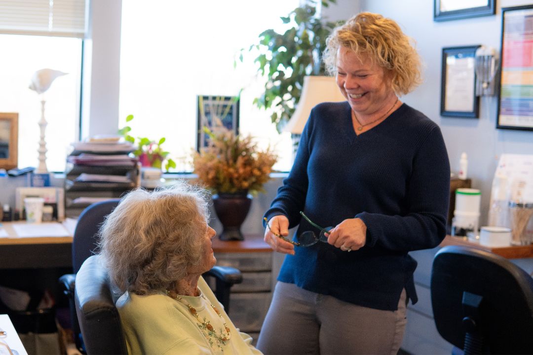 Smiling female audiologist holding glasses stands and talks to an elderly woman seated in an office with plants and paperwork in the background.