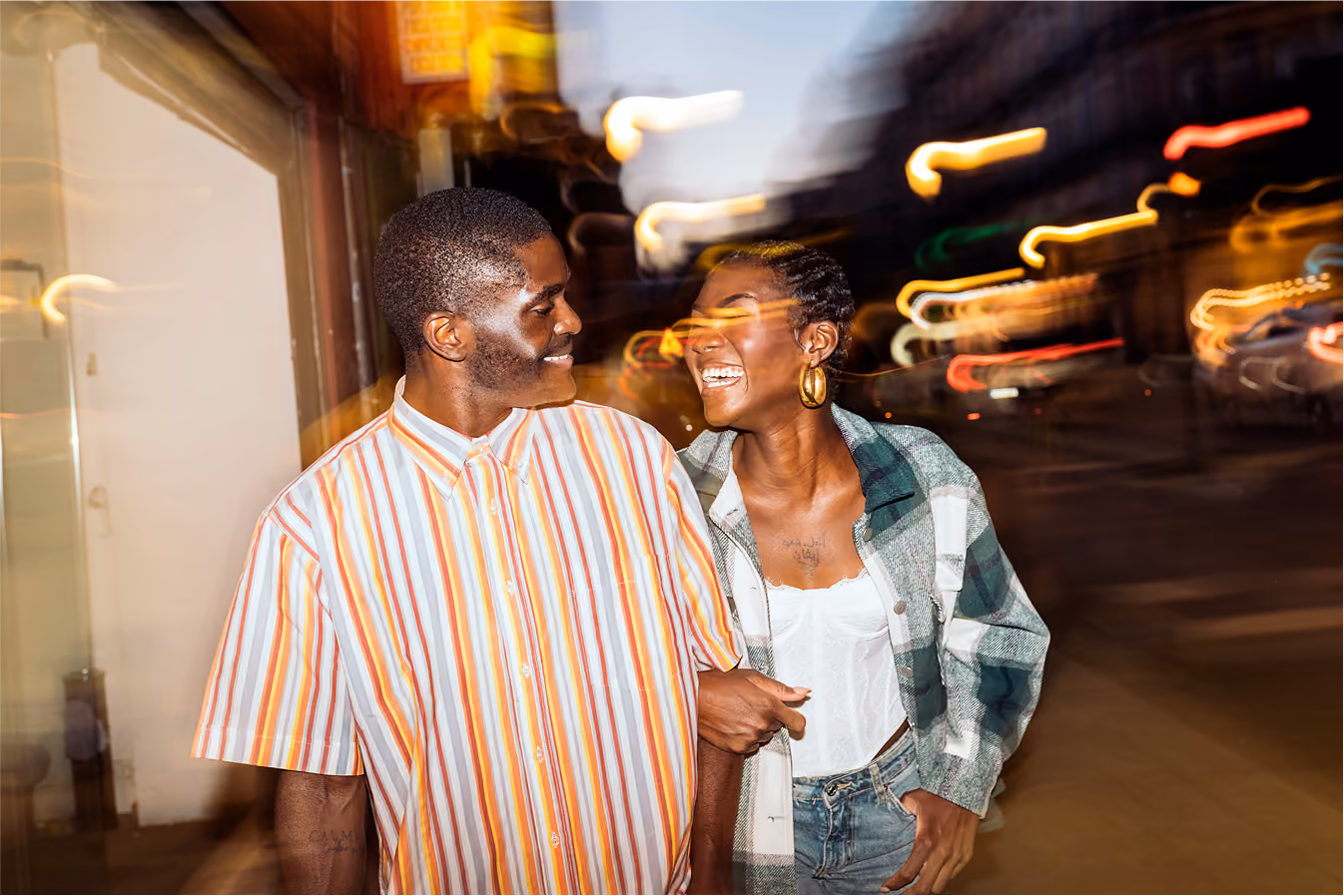 Happy couple walking arm in arm on a city street at night with blurred lights in the background.