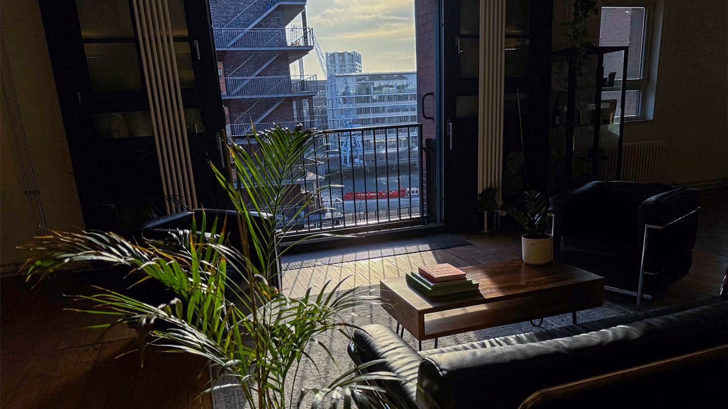 Innenraum mit Sofa, Sesseln, Pflanzen und offenem Fenster mit Blick auf Wasser und Gebäude.