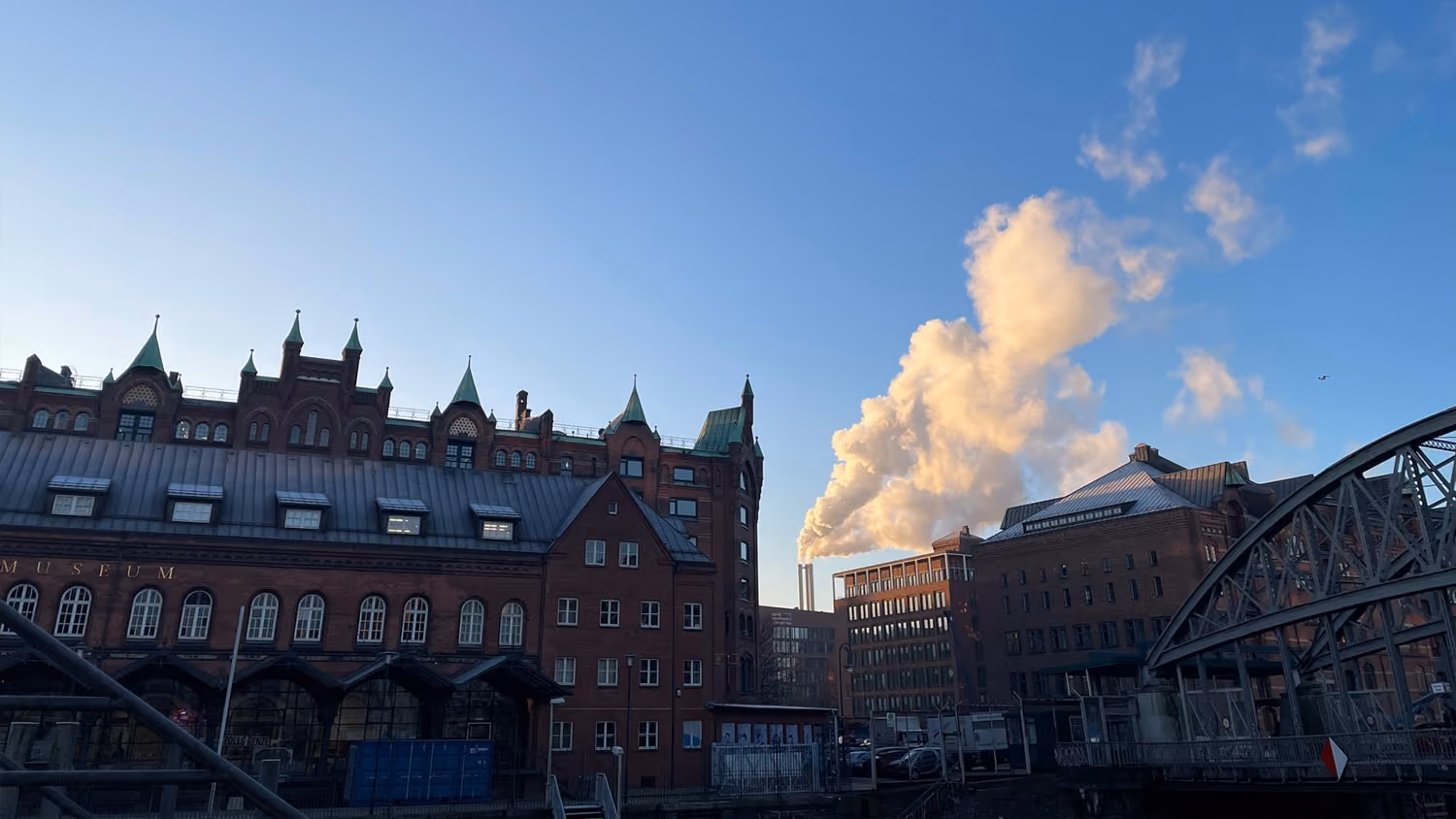 Historische Backsteingebäude mit grünen Dächern neben einem Industriegebäude und Rauch unter klarem Himmel.