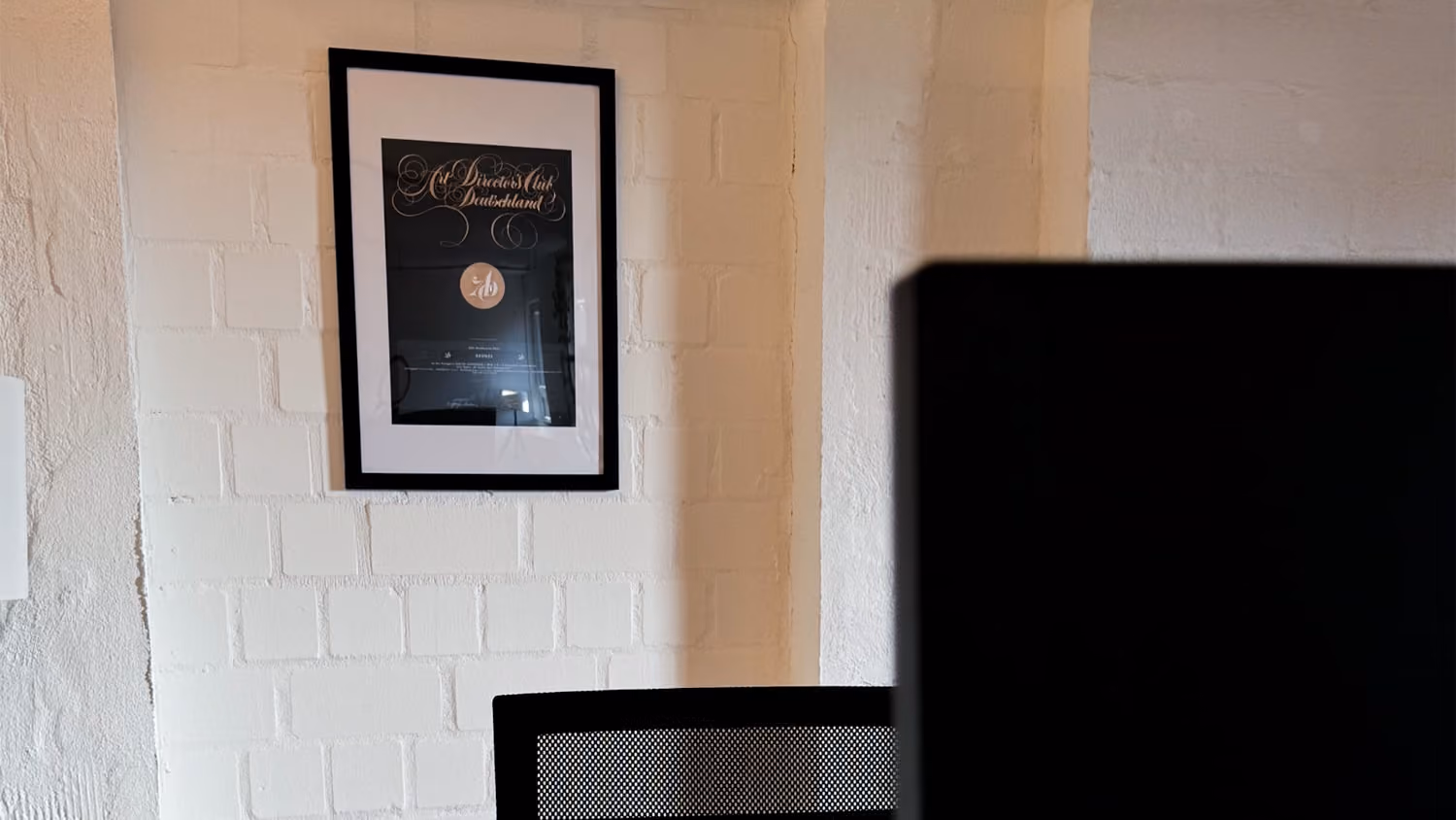 Schwarzes Bild in schwarzem Rahmen an weißer gemauerter Wand über einem schwarzen Stuhl.