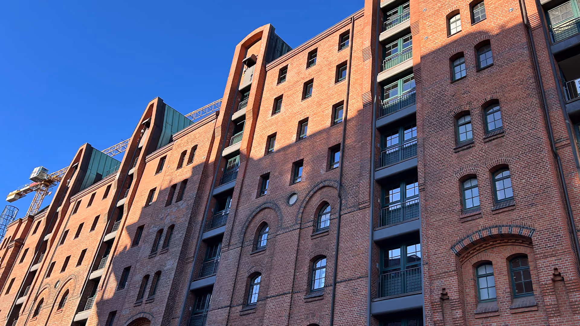 Rote Backstein-Fassade eines mehrstöckigen Gebäudes bei Sonnenschein vor blauem Himmel.
