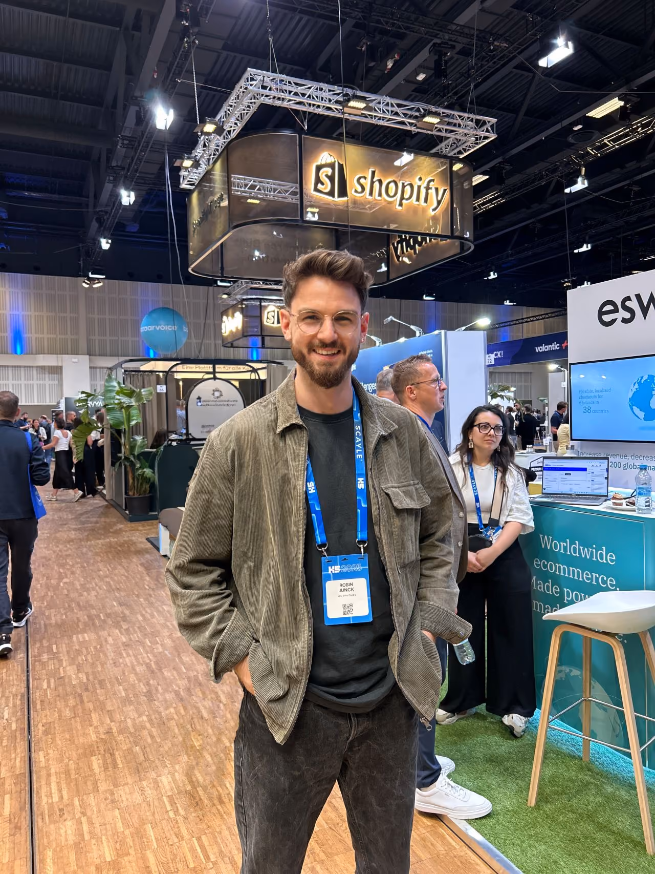 Ein lächelnder Mann mit Brille steht vor einem Shopify-Messestand auf einer Messe.