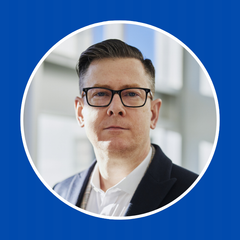Man with short brown hair and glasses wearing a white shirt and dark blazer in a bright office environment.