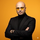 Confident bald man wearing yellow-tinted glasses and a black turtleneck with blazer, standing against an orange background.