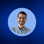 Smiling man with glasses and beard wearing a light blue collared shirt against a blue background.