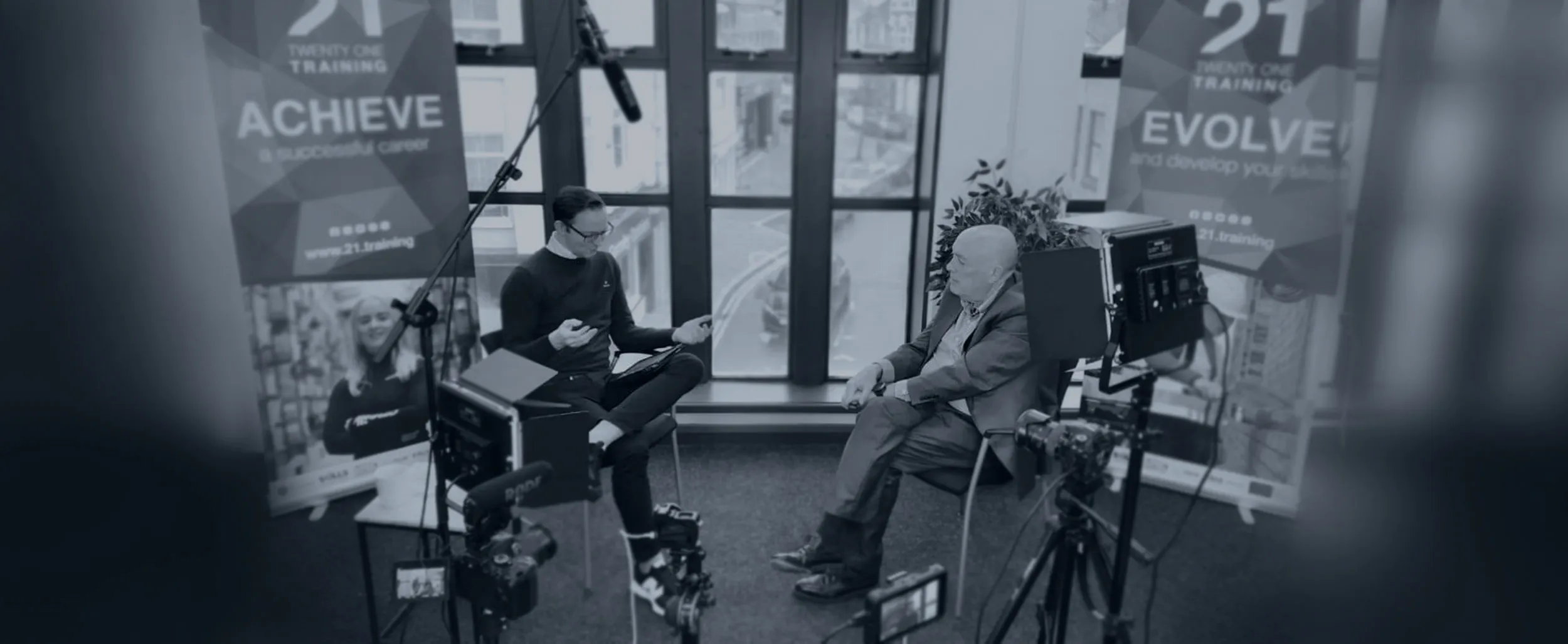 Two men seated and conversing in a studio setup with professional lighting, cameras, and boom microphone in front of large windows.
