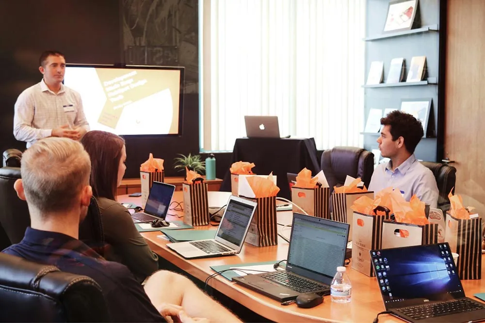 Four people in a conference room with laptops, listening to a man presenting in front of a screen.